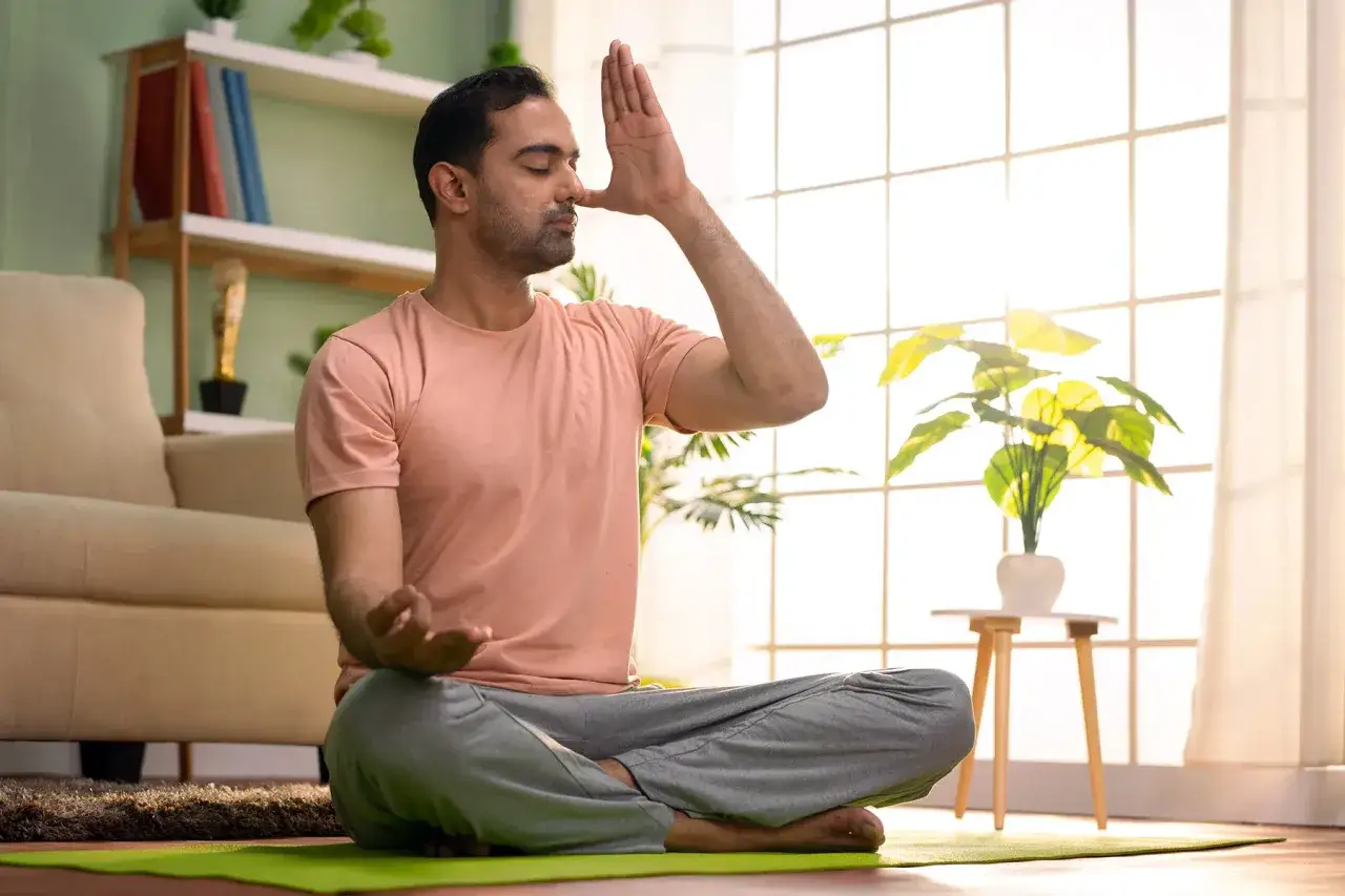 Mężczyzna w domu praktykuje techniki oddychania pranayama, siedząc w pozycji lotosu na macie.