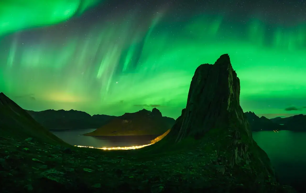 Grüne Aurora Borealis tanzt über einem ruhigen Fjord mit einer kleinen, beleuchteten Stadt am Ufer und dramatischen Bergformationen.