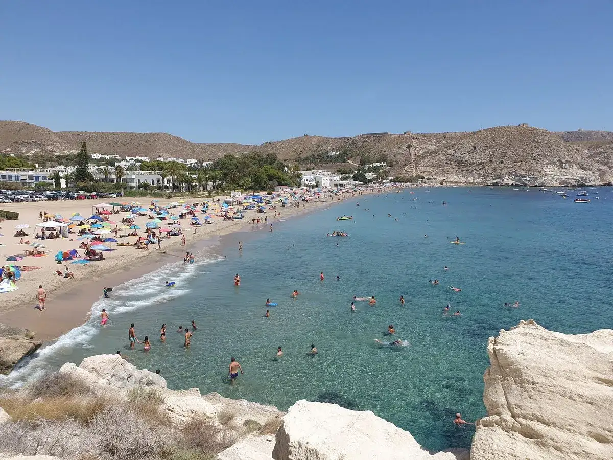 Gente disfrutando en la playa de agua amarga, con sombrillas, bañistas y aguas cristalinas bajo un cielo azul.