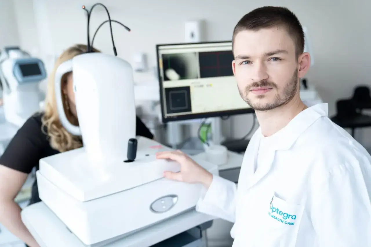 Okulista bada pacjentkę z zaćmą wikłającą. Mężczyzna w białym fartuchu przy aparaturze diagnostycznej.
