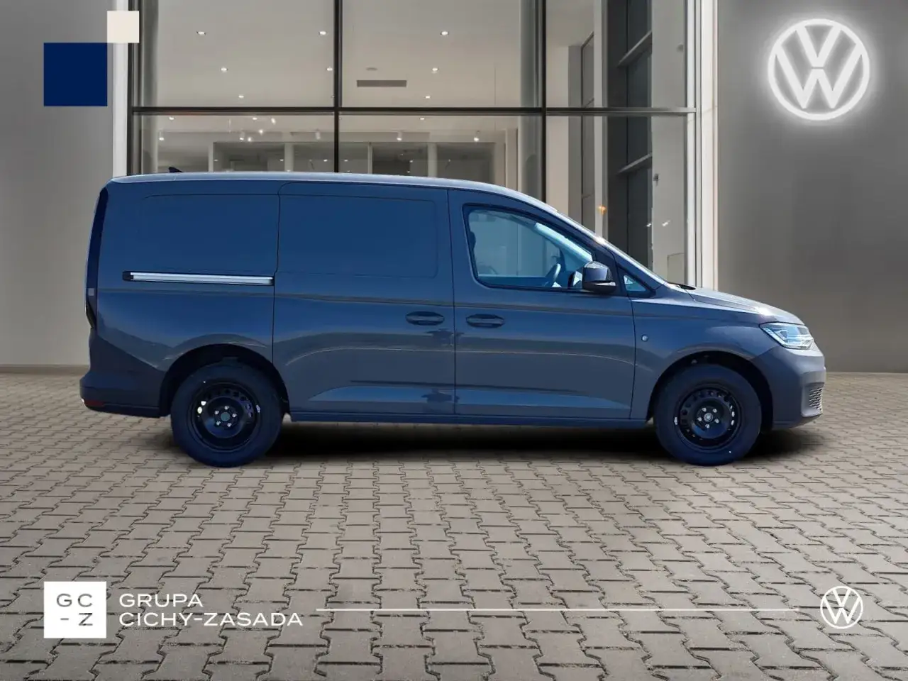 Ciemnoszary Volkswagen Caddy 3, zaparkowany przed salonem z podświetlanym logo marki.