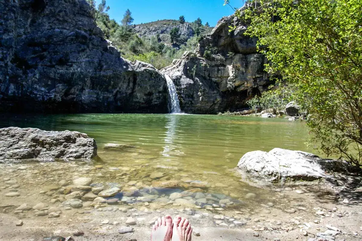 Pies descalzos en la orilla del **barranco de la Encantada**, con una cascada cayendo en una poza de agua verde rodeada de rocas y vegetación.