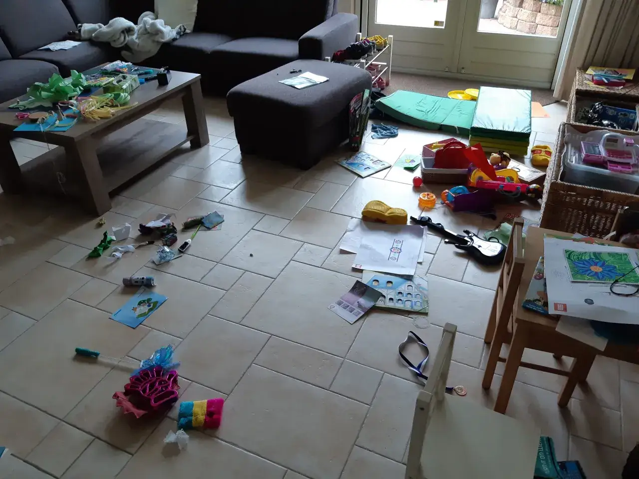 Een rommelige woonkamer met speelgoed en knutselspullen verspreid over de vloer. Een duidelijke