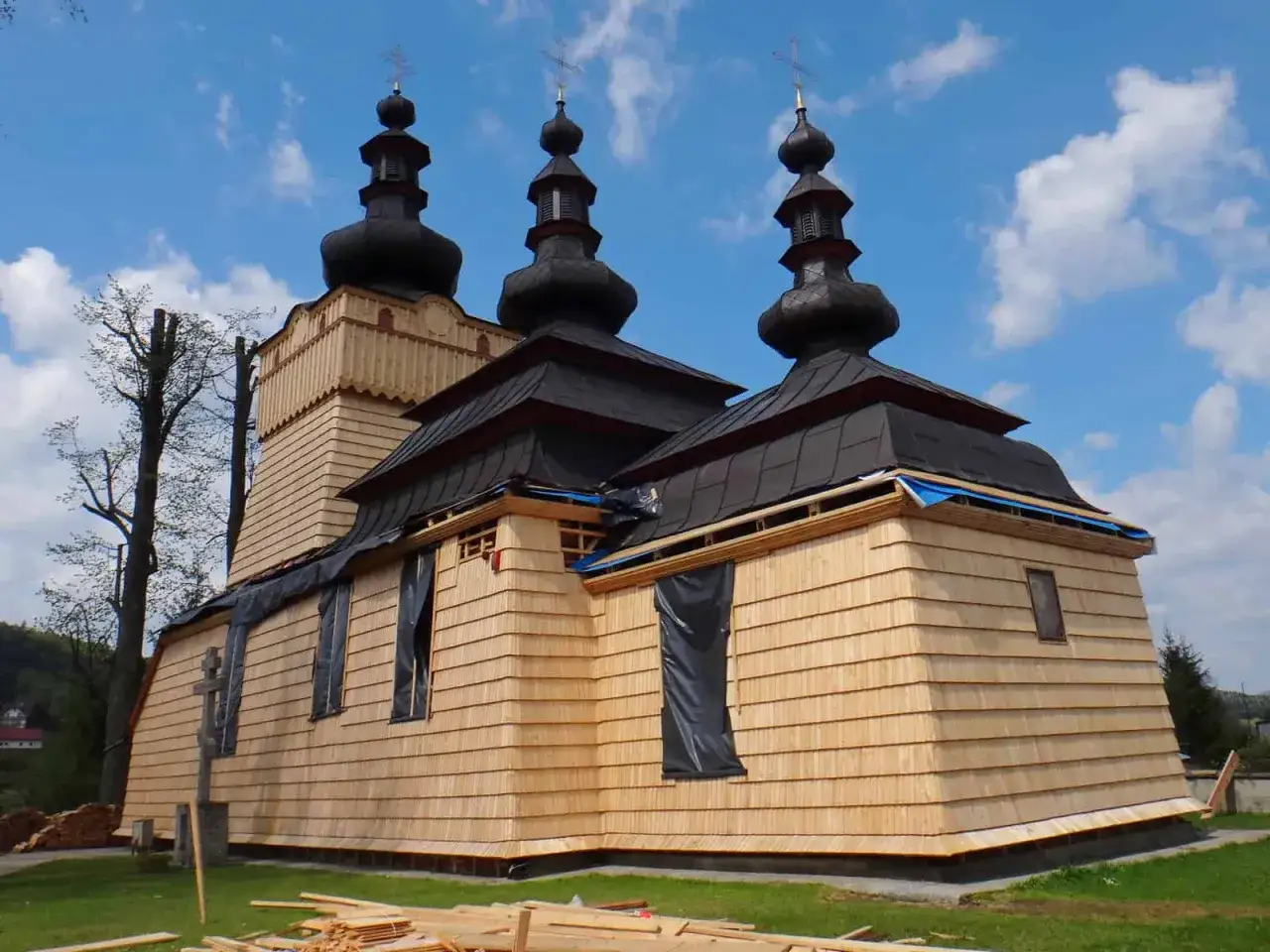 Drewniana cerkiew z kopułami, w trakcie remontu, na tle błękitnego nieba z chmurami. W oddali widać zalesioną g&oacute;rę Jawor.