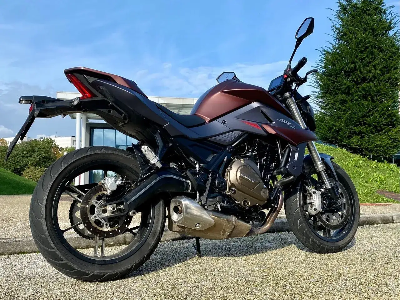 Moto SRK 700 color bronzo e nero, parcheggiata su un vialetto ghiaioso con un edificio moderno e un albero sullo sfondo.