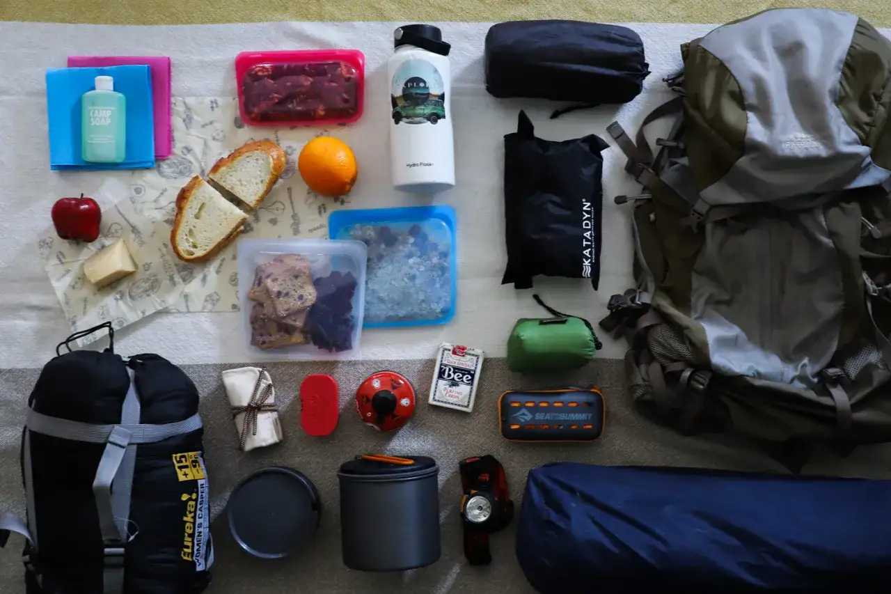 Proviant für eine Wanderung: Rucksack, Schlafsack, Kochgeschirr, Wasserflasche, Obst, Brot, Kekse und Fleisch.