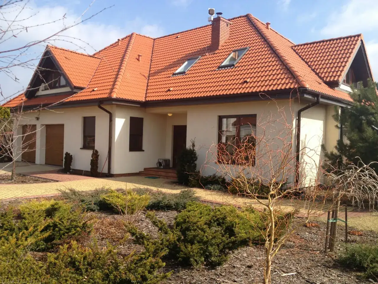 Nowoczesny dom z ceglastym dachem i jasną, szarą elewacją, otoczony zielenią i brukowanym podjazdem.