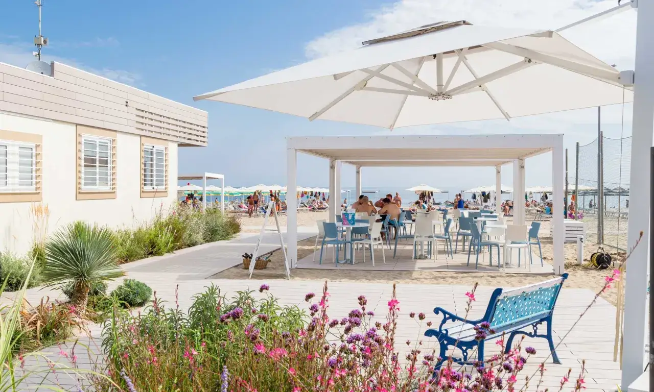 Un angolo di relax nella darsena di Riccione: tavoli all'aperto sotto un pergolato bianco, con la spiaggia e il mare sullo sfondo.