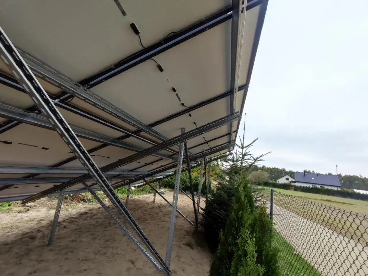 Instalacja fotowoltaiczna w granicy działki, widok od spodu paneli. Metalowa konstrukcja wspiera panele, obok rosną drzewka.