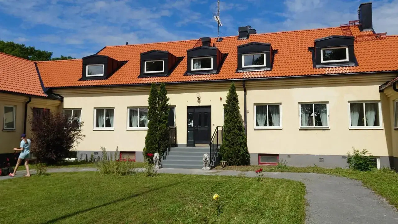 Gult vandrarhem på Gotland med orange tak och gröna träd framför. En person klipper gräsmattan.
