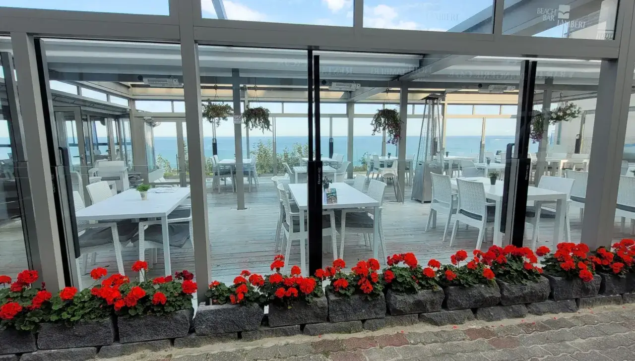 Letnia, przeszklona restauracja z widokiem na morze. Stoliki i krzesła w hotelu Lambert, przed nimi czerwone pelargonie.