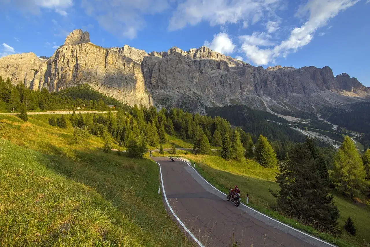 Motociclisti sfrecciano sulla strada panoramica del Sella Ciampigotto, tra prati verdi e imponenti vette rocciose sotto un cielo azzurro.