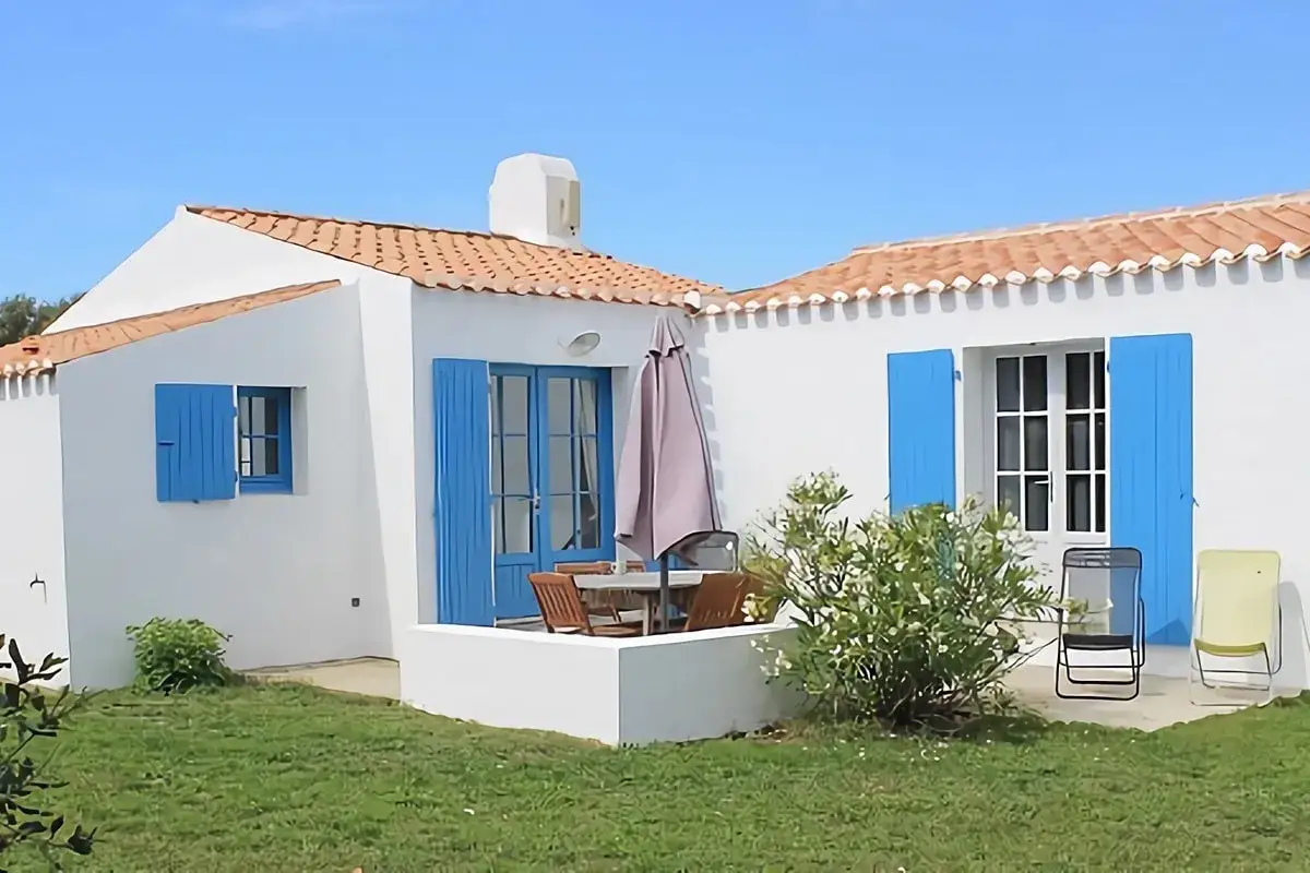 Charmante maison vendéenne blanche aux volets bleus vifs, avec terrasse et jardin verdoyant sous un ciel clair.