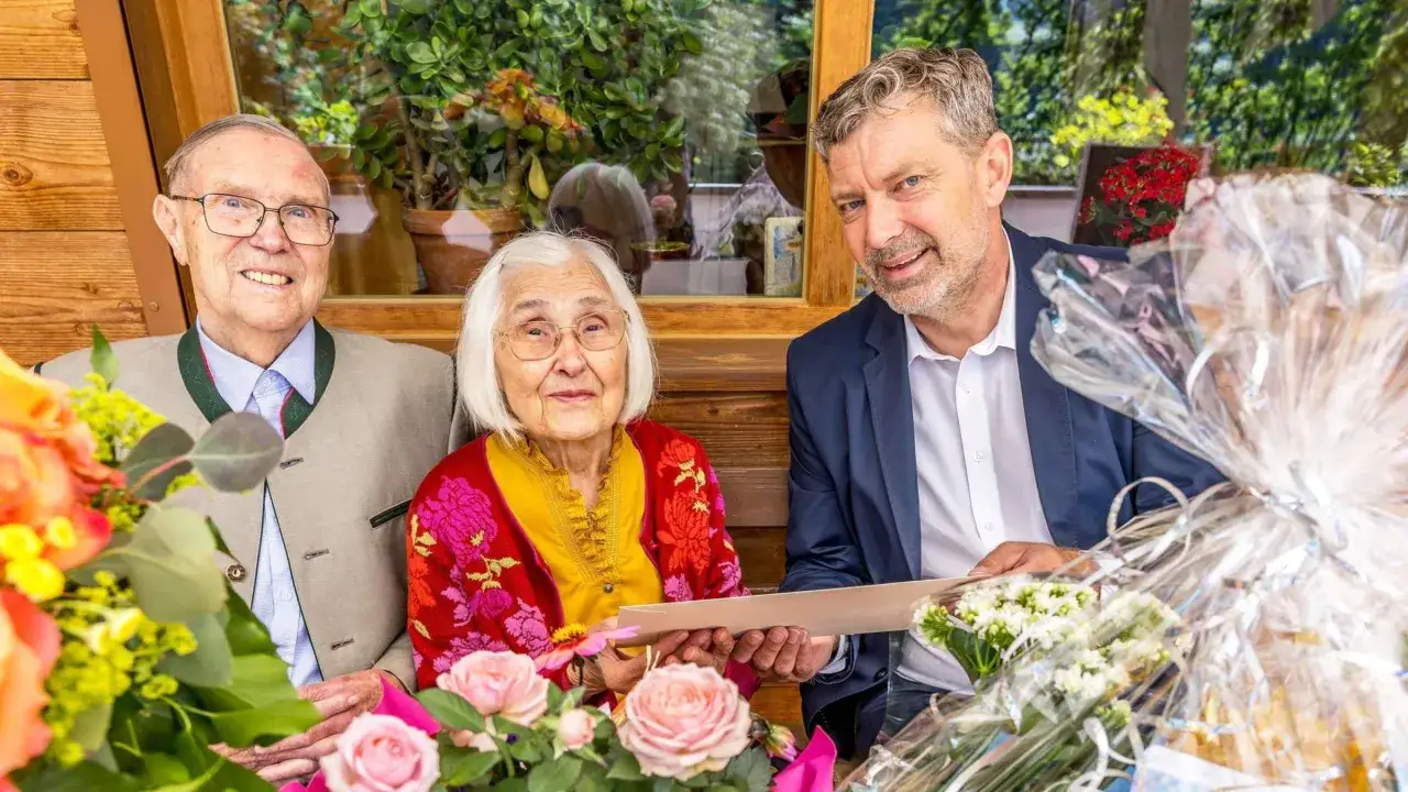 Ein älteres Paar feiert Gnadenhochzeit, wieviel Jahre sie wohl verheiratet sind? Sie sitzen mit Blumen und Geschenken im Freien.