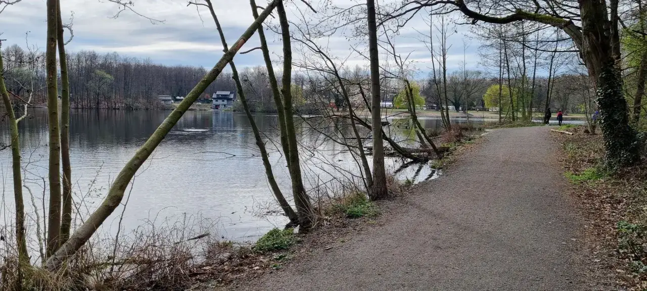 Ein Weg lädt zum spazieren gehen am See in NRW ein. Bäume säumen das Ufer, im Hintergrund Häuser und Campingwagen.