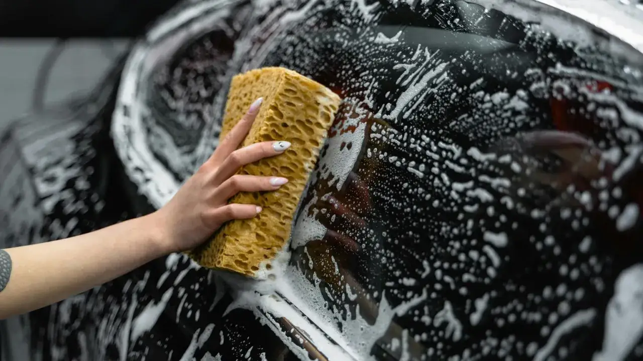Ręka z gąbką myje czarne auto pokryte pianą. Gdy brakuje szamponu, gąbka może pom&oacute;c w czyszczeniu.
