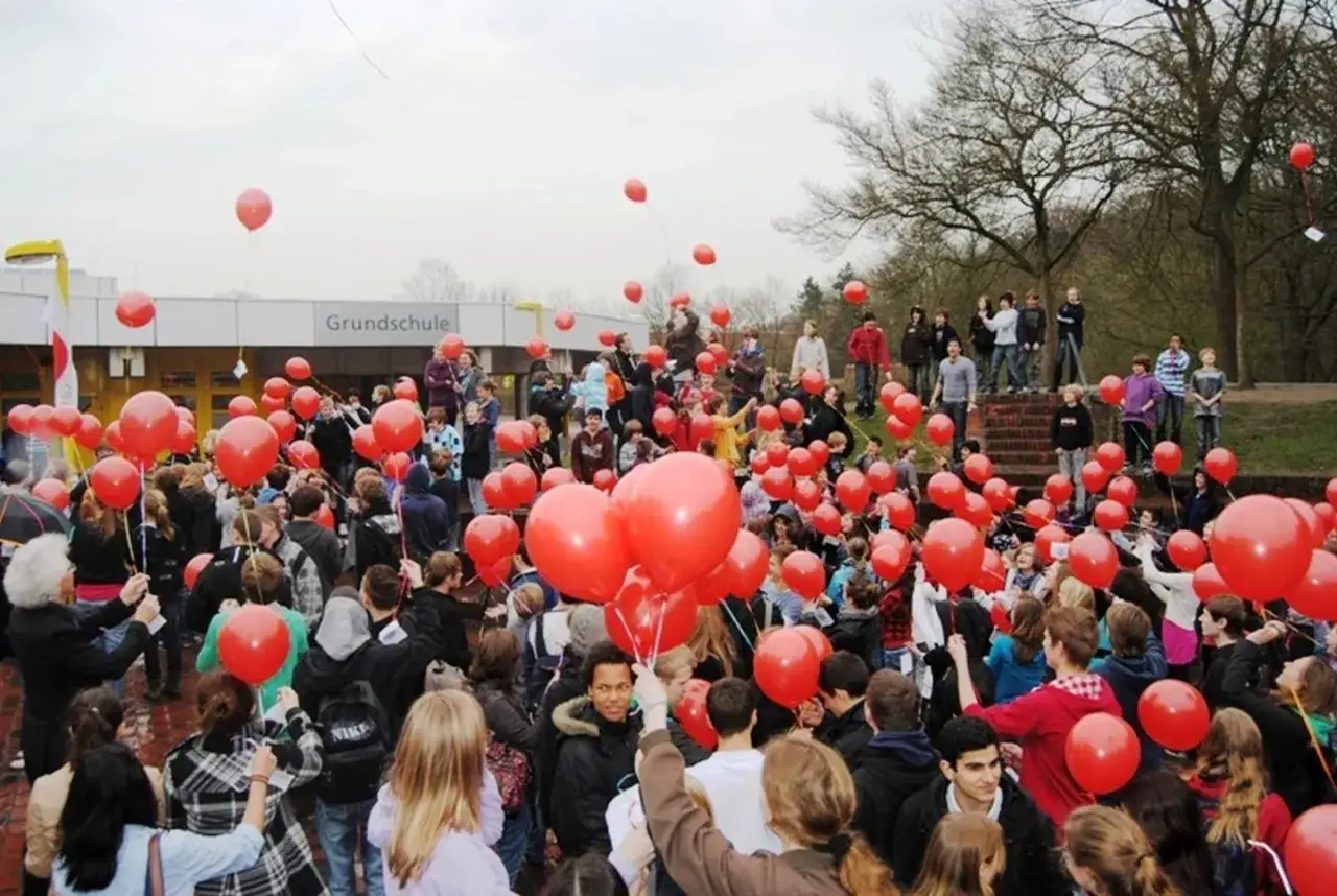 Eine große Gruppe von Menschen, darunter viele Kinder, hält rote Ballons vor einer Grundschule. Ein Zeichen der Solidarität.