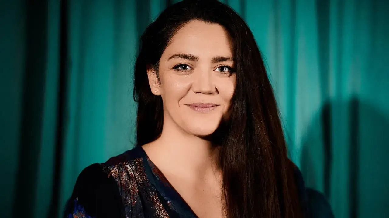 Jenny Nordlander ler mot kameran, med mörkt hår som faller över axeln. Bakgrunden är en turkos gardin.
