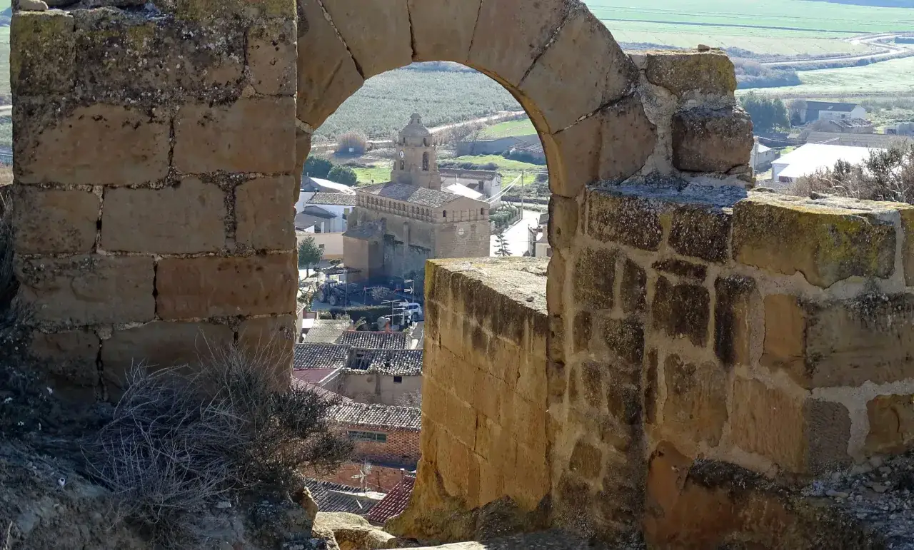 Vista de un pueblo a través de un arco de piedra. Se ve una iglesia con torre y tejados de teja. El paisaje circundante es verde y rural, con campos y vegetación. La alberuela de tubo se integra en la arquitectura antigua.