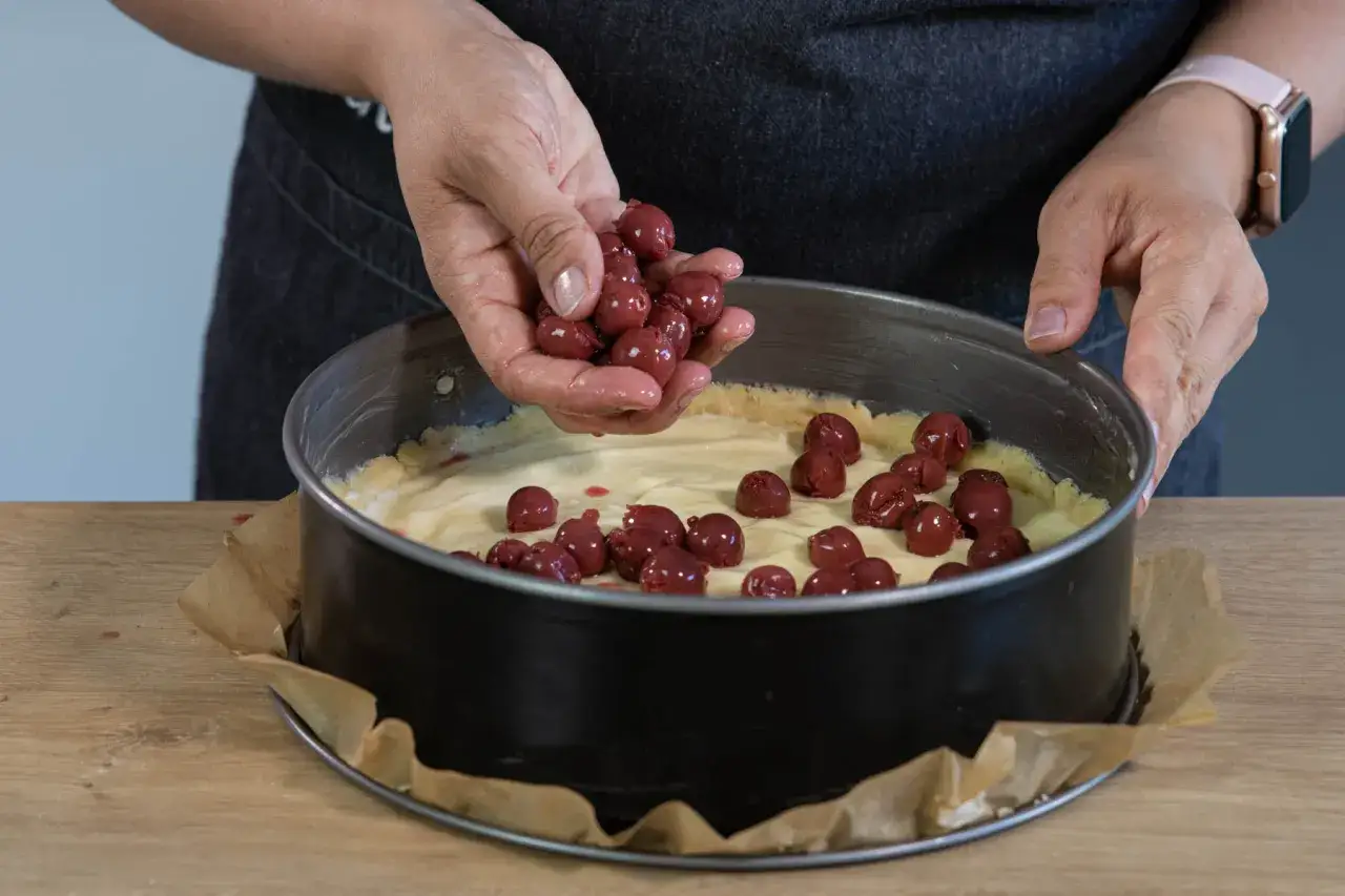 Hände legen Kirschen auf eine cremige Füllung in einer Springform. Bald entsteht ein köstlicher Kirschkuchen mit Pudding.