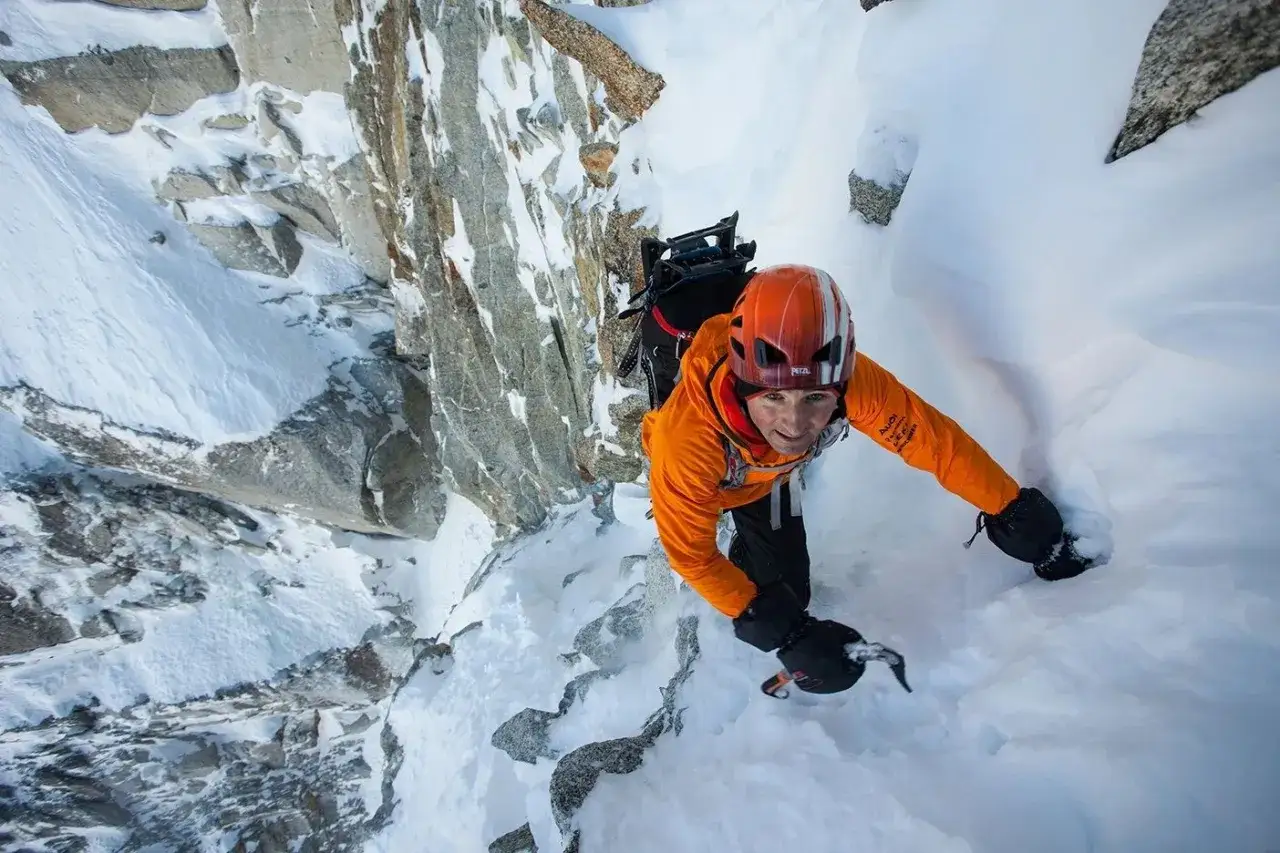 Ueli Steck klettert in orangefarbener Jacke und Helm einen eisigen, schneebedeckten Felsen hinauf.