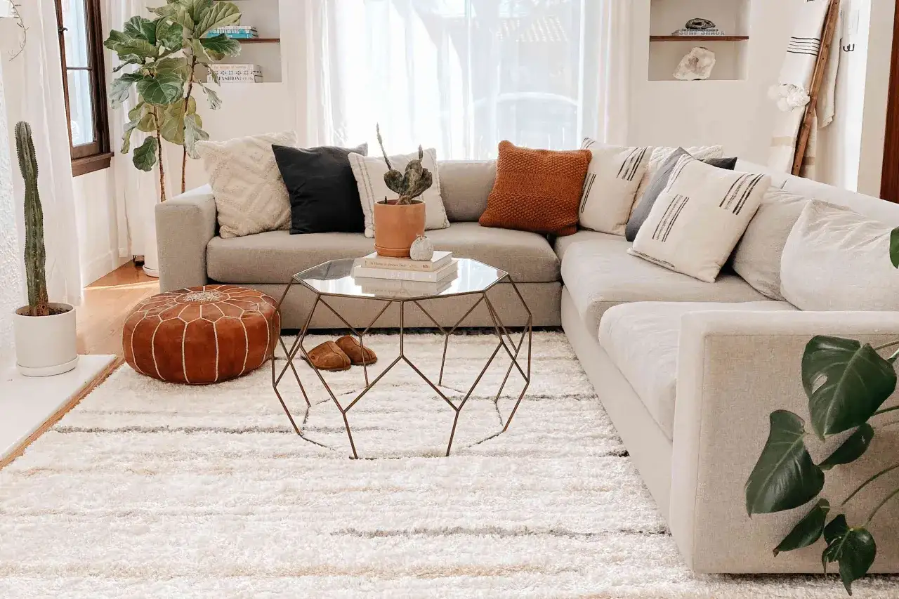 Ein flauschiger, weißer **Teppich unter dem Sofa** schafft Gemütlichkeit. Darauf ein brauner Pouf und ein moderner Couchtisch.
