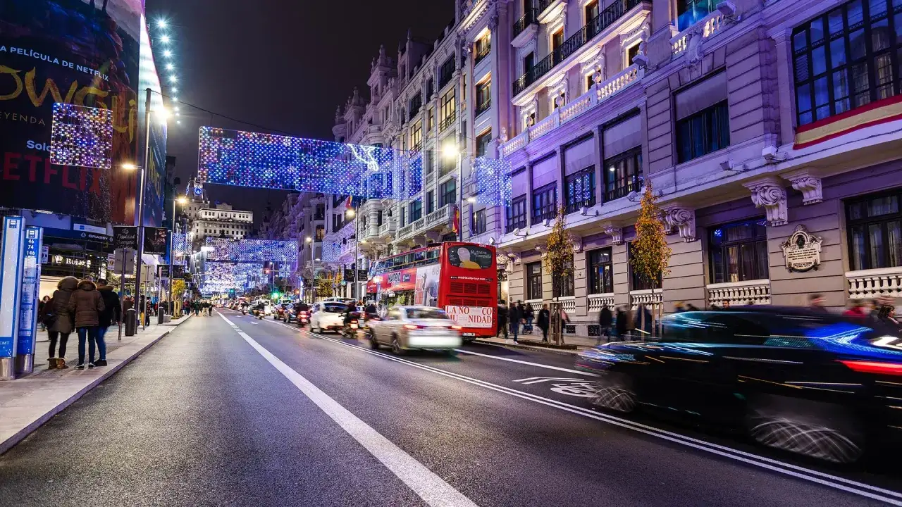 Nocny Madryt w lutym, rozświetlony świątecznymi iluminacjami. Ruch uliczny, czerwony piętrowy autobus i ludzie spacerujący po chodniku.