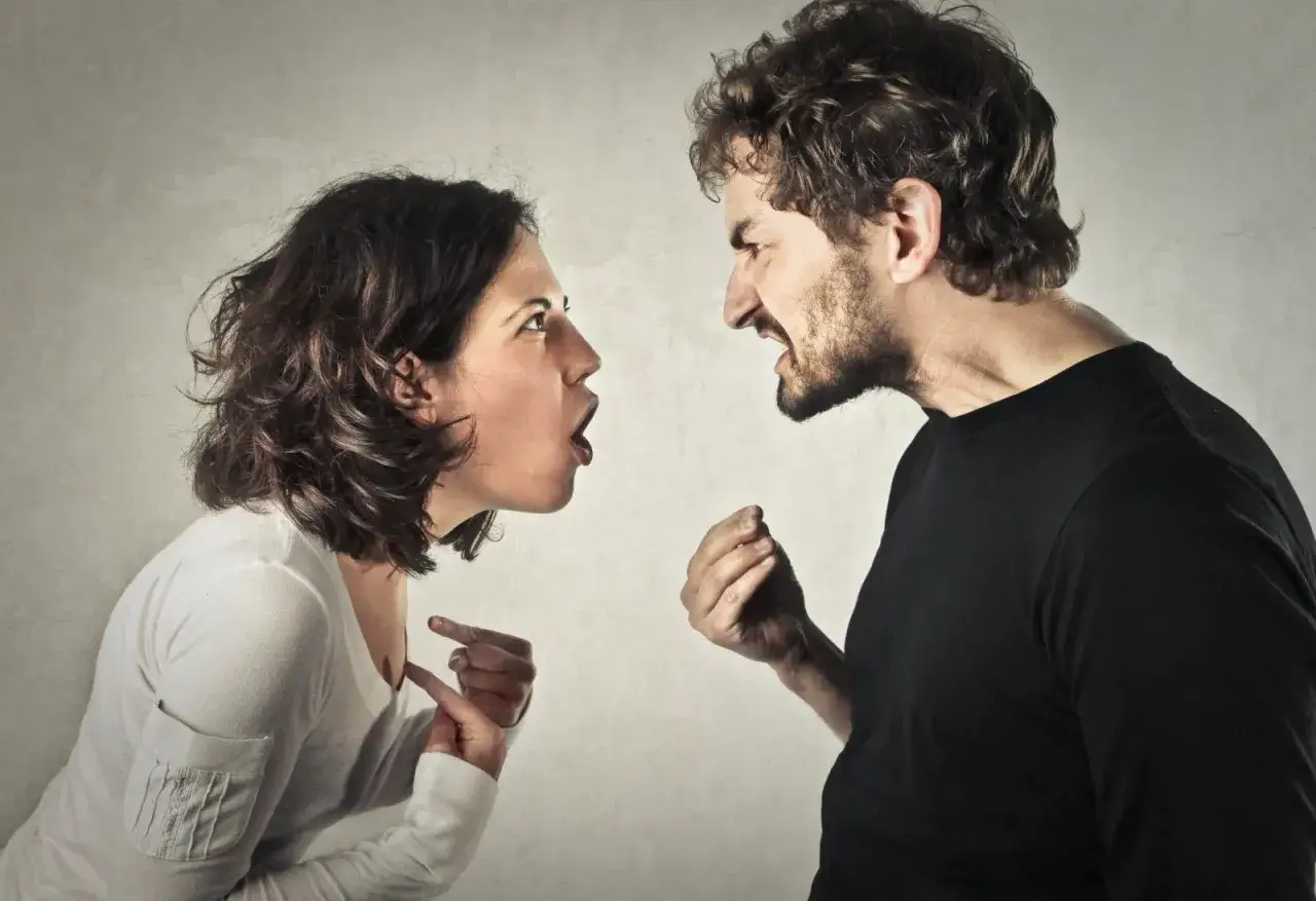 Un homme en colère crie sur une femme, qui semble être victime d'une femme manipulatrice. Leurs visages sont proches, exprimant une forte tension.