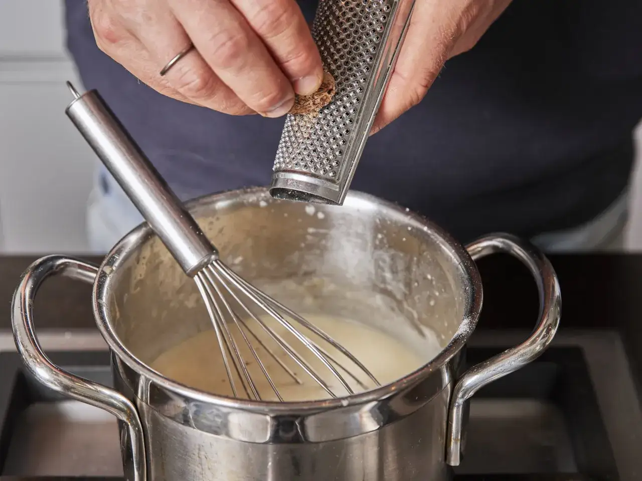 Eine Hand reibt Muskatnuss &uuml;ber einen Topf mit k&ouml;chelnder B&eacute;chamelsauce, die mit einem Schneebesen verfeinert wird.