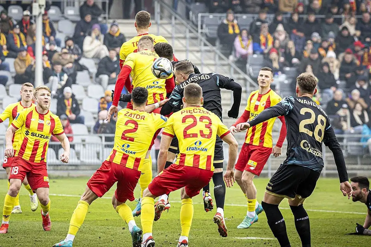 Piłkarze w żółto-czerwonych strojach walczą o piłkę w powietrzu. W tle trybuny kibiców. Przebieg: Pogoń Szczecin – Korona Kielce.