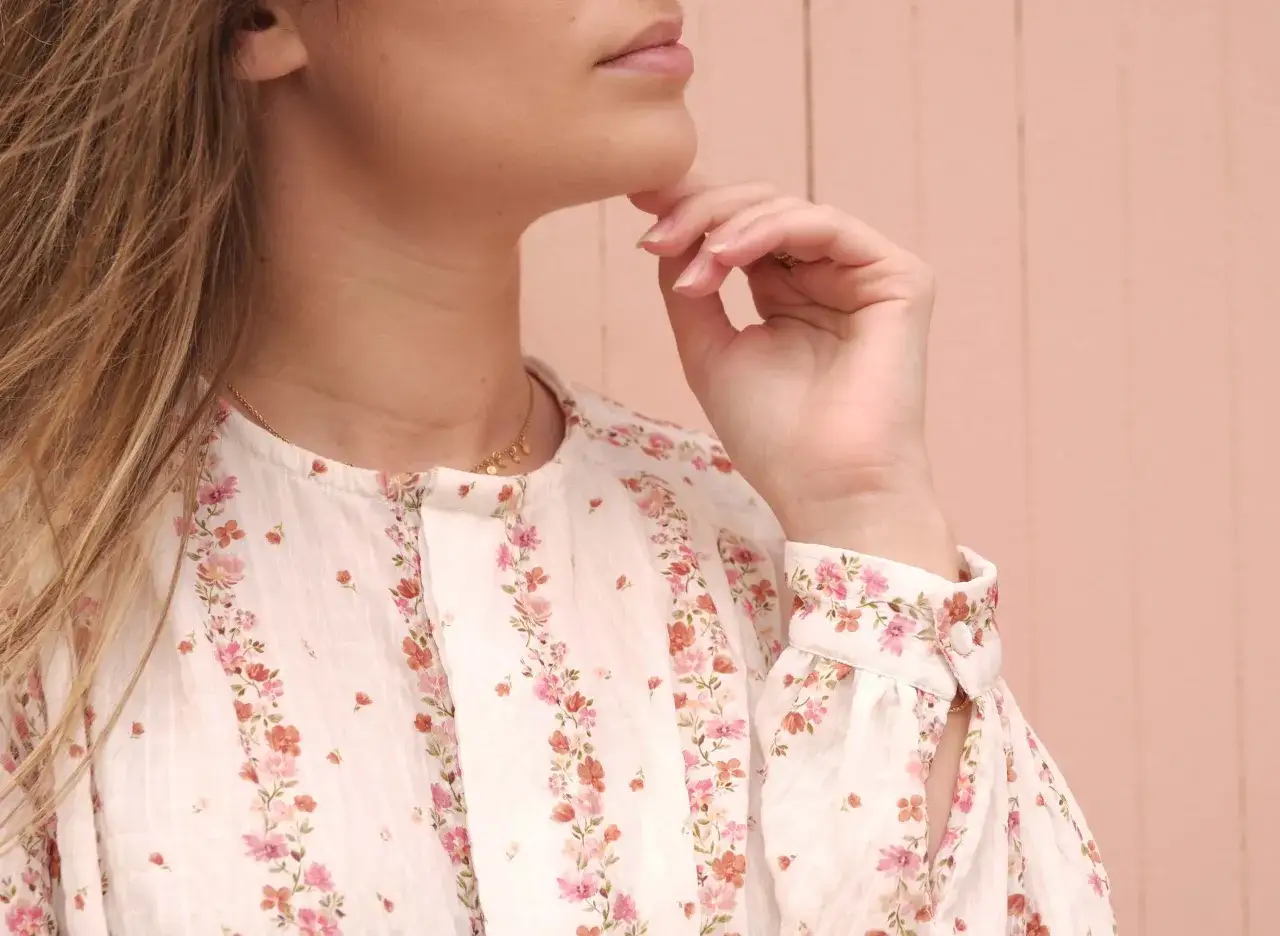 Gros plan sur le boutonnage femme d'une blouse &agrave; fleurs roses et corail, avec une main pos&eacute;e sur le menton.
