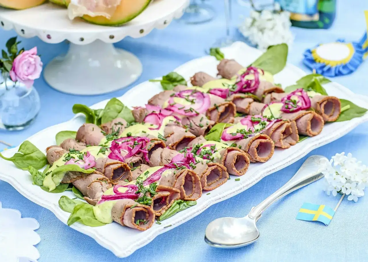 Rullad rostbiff med grön sås och rödlök, perfekt som enkel buffémat till många. Serveras på en vit bricka med gröna blad.