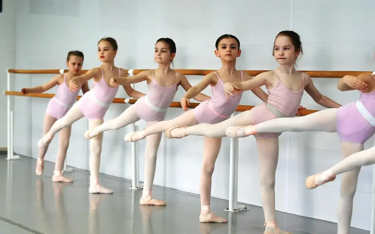 Bambine in tutù rosa praticano la **danza propedeutica** alla sbarra, con le gambe tese e i piedi in punta.