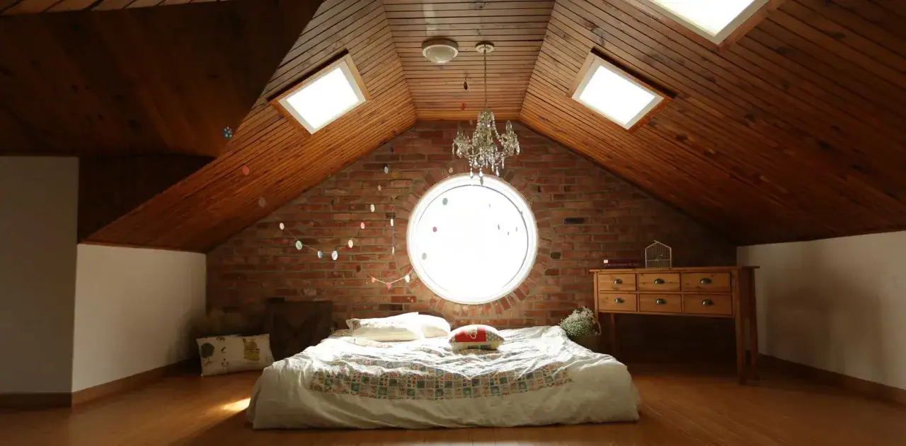 Camera puntata verso un letto in mansarda, con il soffitto in legno e la sua evidente pendenza tetto. Finestre sul tetto e un oblò circolare illuminano la stanza.