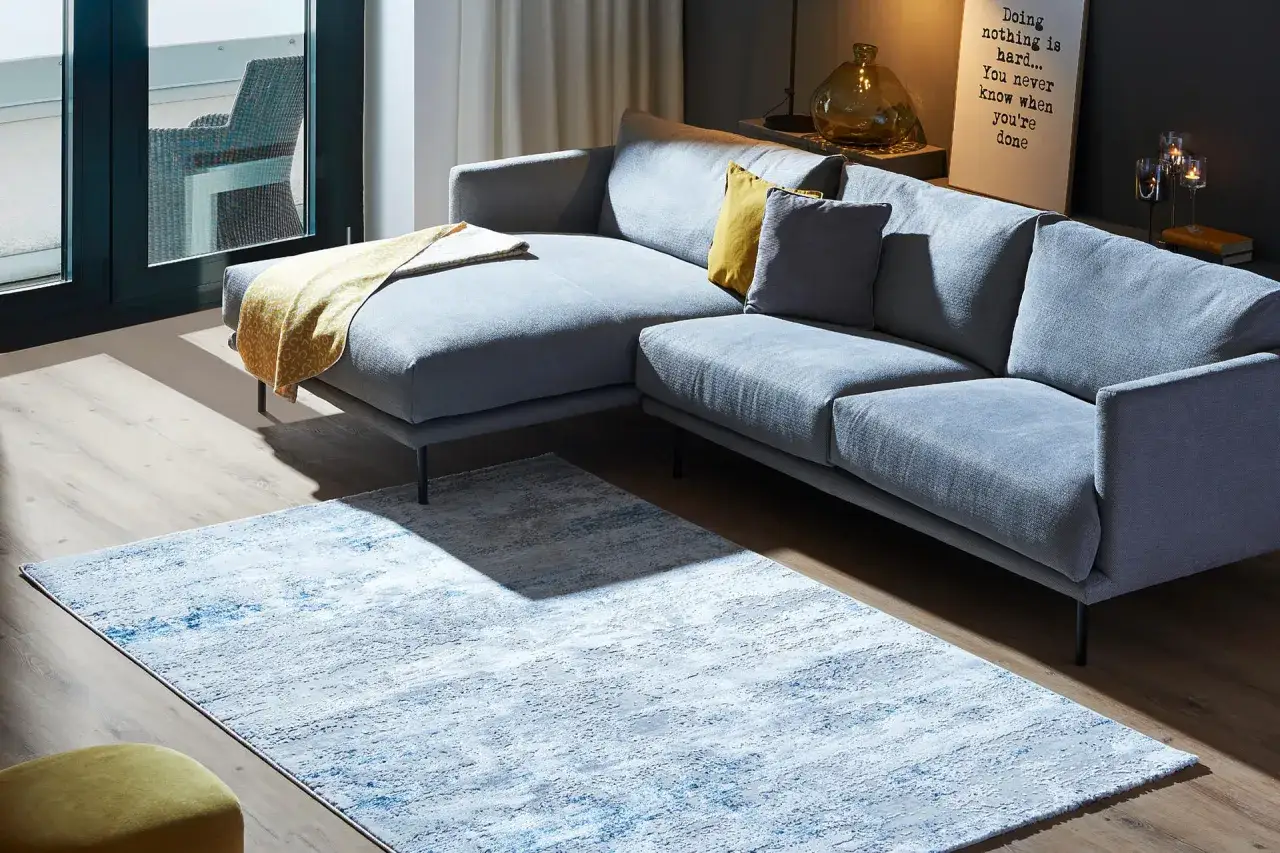 Ein moderner, grauer Teppich liegt unter dem Sofa und davor, mit einem Hauch von Blau und Wei&szlig;.