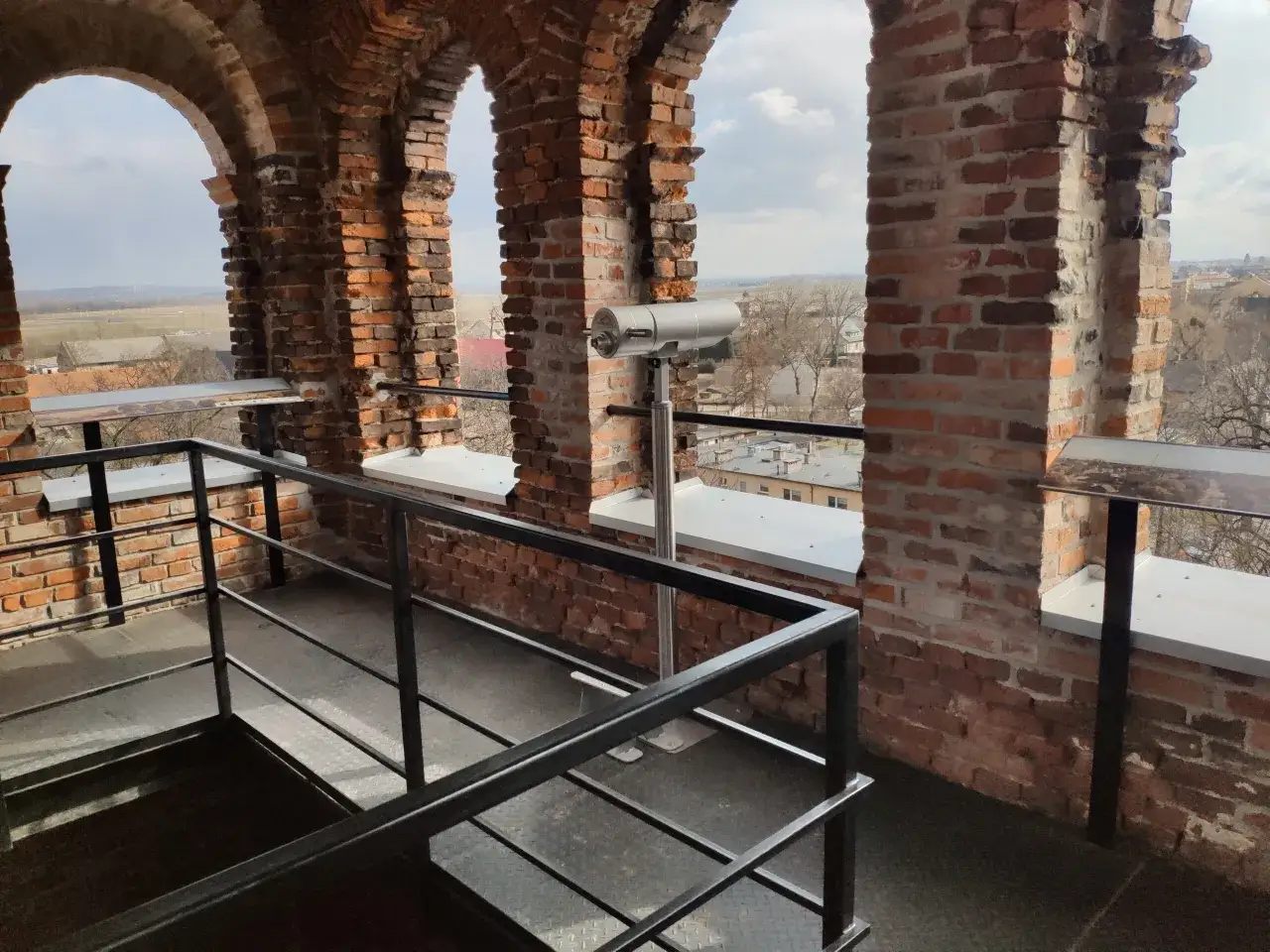 Widok z wieży widokowej na zamek w Tworkowie. Metalowa balustrada i luneta turystyczna na tle ceglanych arkad i krajobrazu.