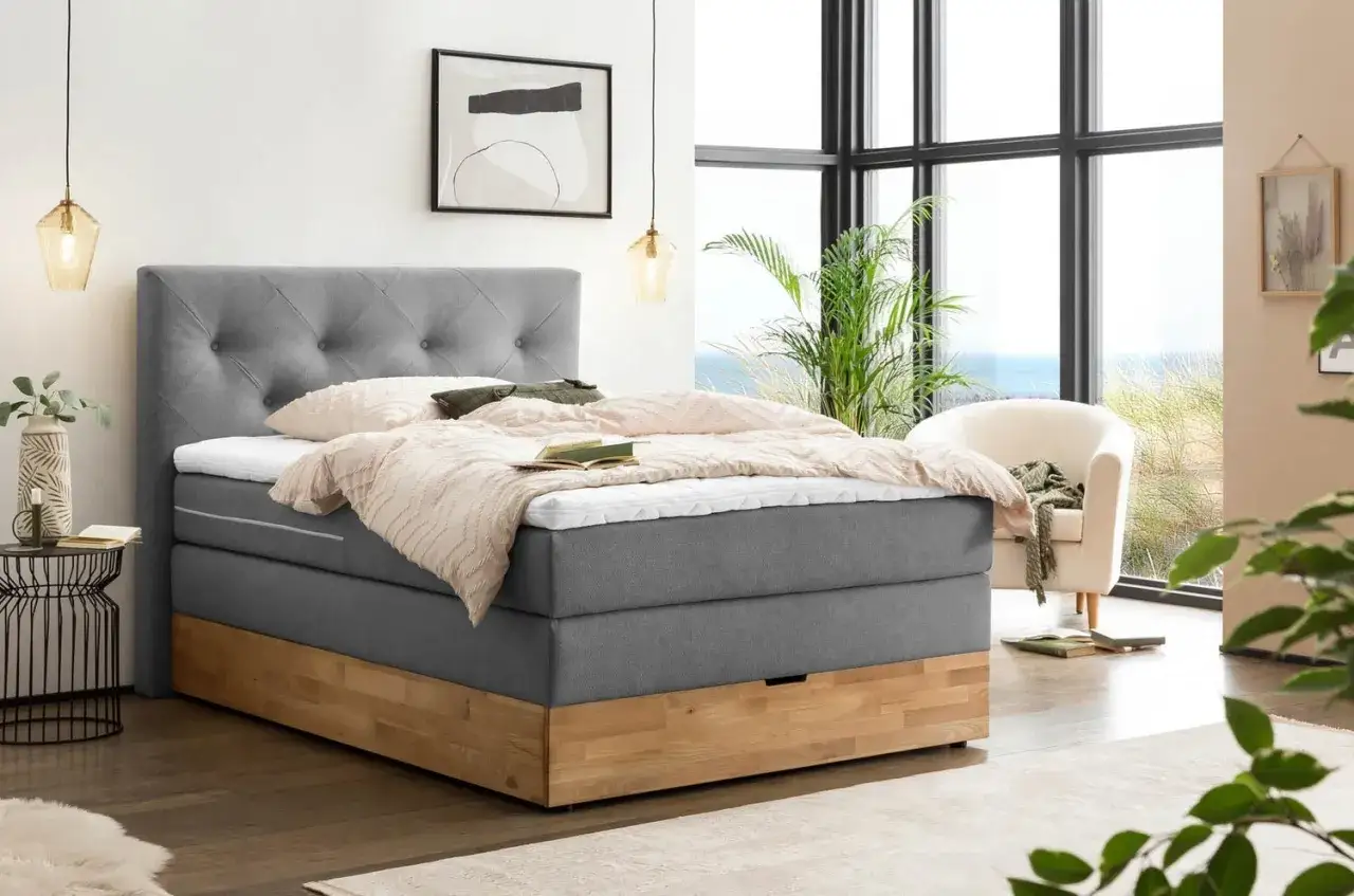 Ein modernes Boxspringbett mit grauem Stoffbezug und Holzsockel steht neben einem gro&szlig;en Fenster mit Meerblick.