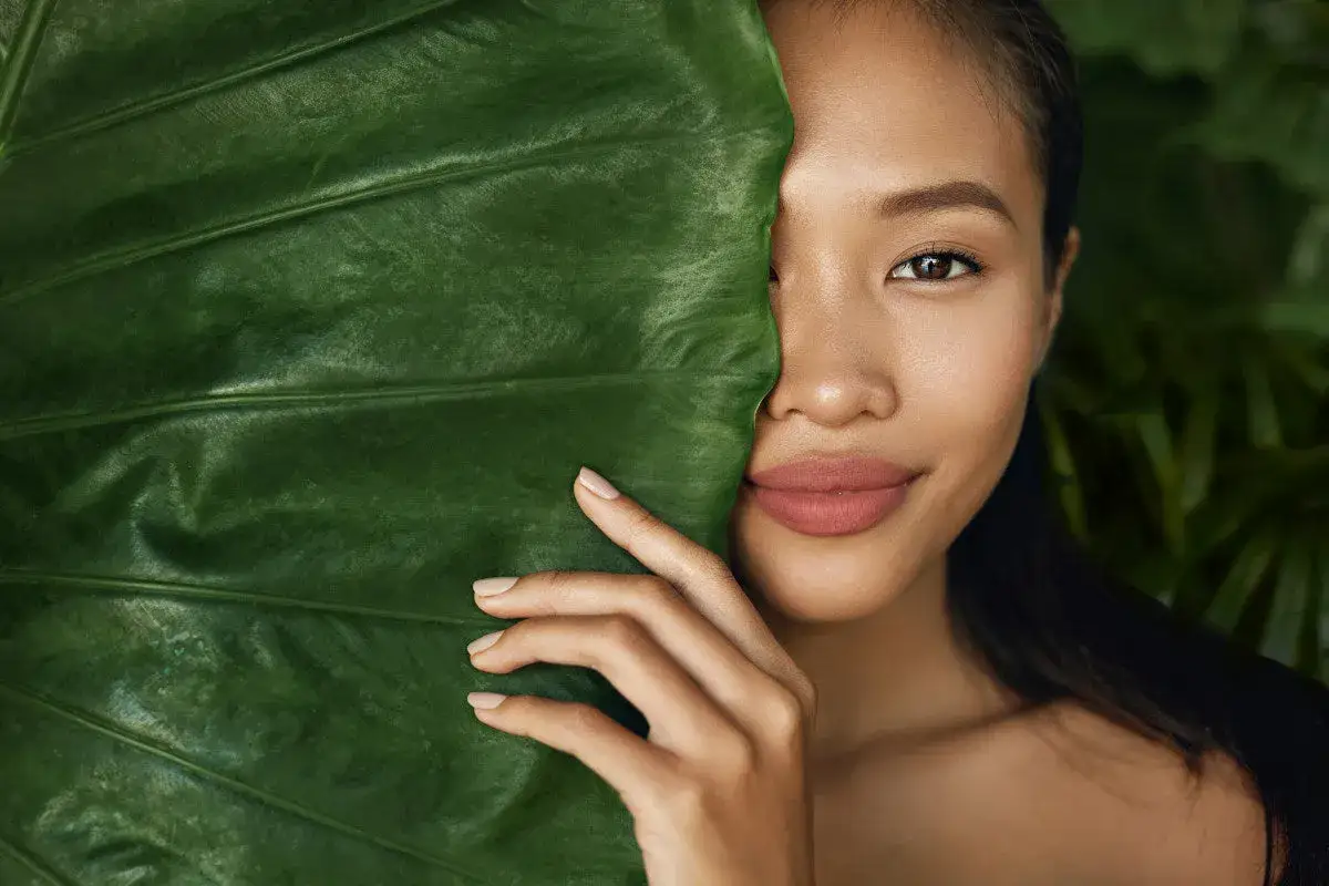 En kvinna med naturlig makeup tittar fram bakom ett stort grönt blad.