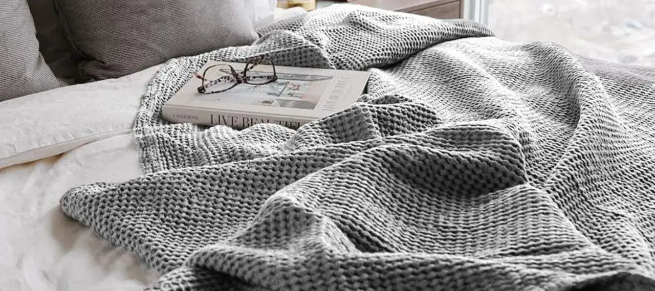 Gem&uuml;tliches Bett mit einer gestrickten Decke, einem Buch und einer Brille. Die Decke ist aus einem weichen Material, das an Perkal erinnert, und l&auml;dt zum Entspannen ein.