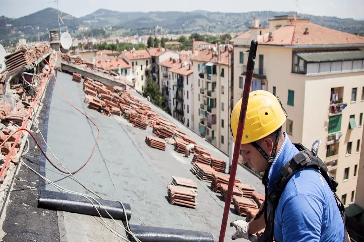 Operaio con casco giallo impegnato nella riparazione tetto casa, con tegole sparse e materiali edili sul tetto di un edificio in citt&agrave;.