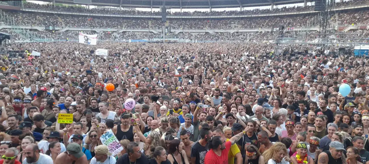 Folla immensa a un concerto, gente esultante con le braccia al cielo. Un cartello recita