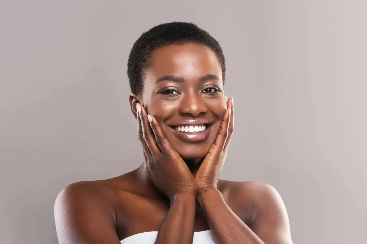 Une femme souriante caresse son visage, sa peau éclatante témoigne d'une hydratation en profondeur.