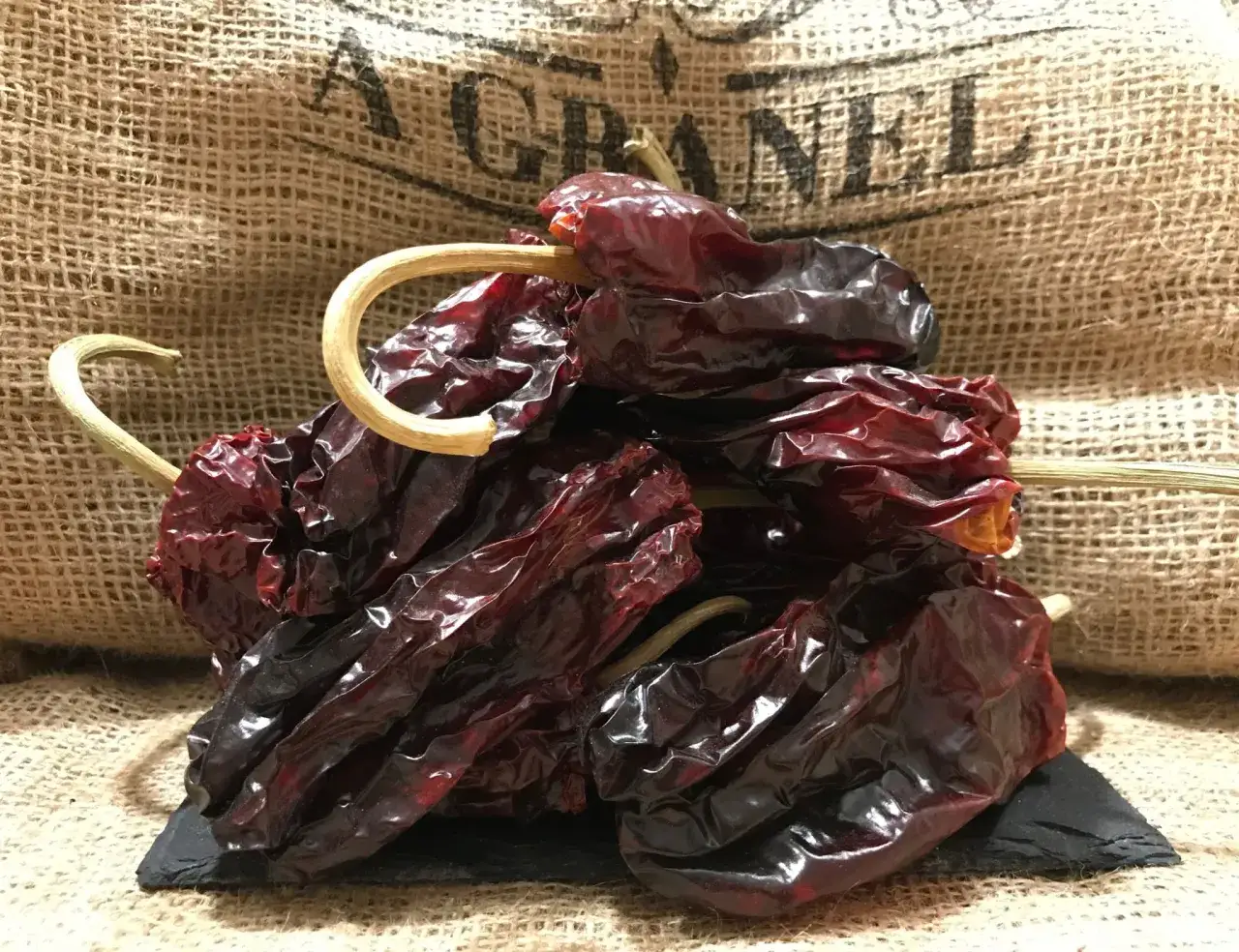 Un manojo de pimientos choriceros secos, oscuros y arrugados, con tallos amarillentos, sobre una pizarra negra y un fondo de arpillera.
