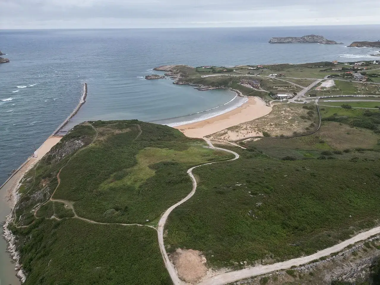 Vista aérea de la **playa de Cuchía**, con su arena dorada, el mar azul y un camino serpenteante entre la vegetación.