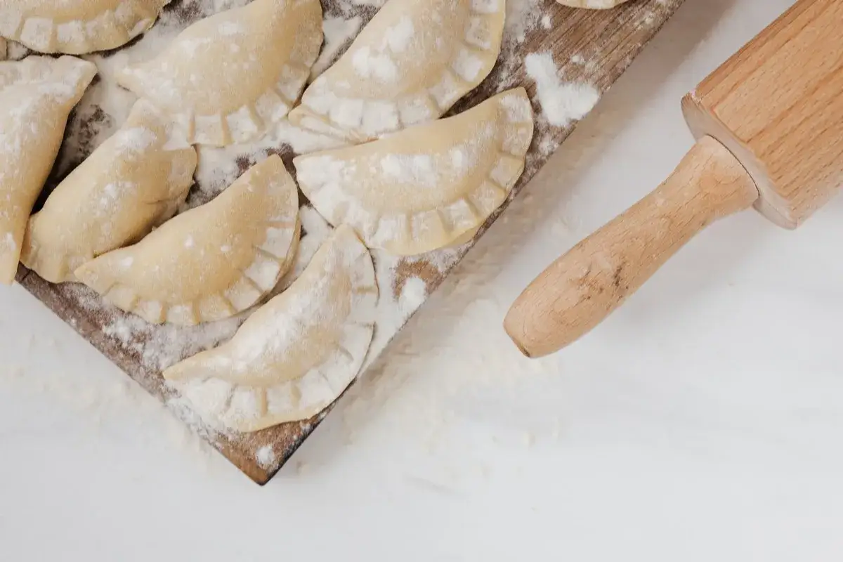 Surowe pierogi na desce, posypane mąką, obok wałek. Idealne do zamrożenia, by wiedzieć, jak przechować ciasto na pierogi.