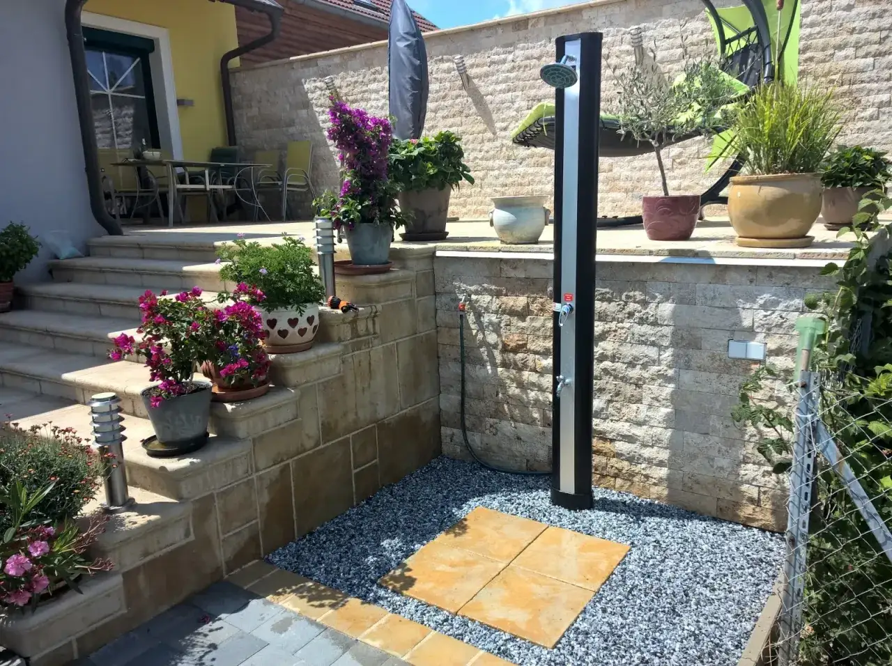 Eine moderne Gartendusche, die man leicht selber bauen kann, steht auf einem Kiesbett mit Steinplatten. Blumen und Pflanzen schmücken die Terrasse.