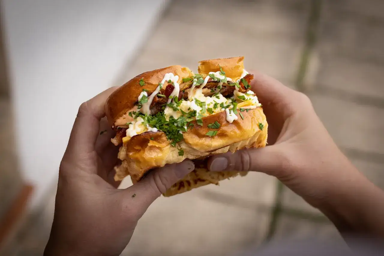 Ein saftiges Egg Drop Sandwich mit Rührei, Speck und Kräutern, gehalten von zwei Händen.