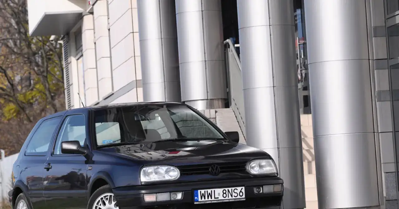 Ciemny Volkswagen Golf III, pierwszy samoch&oacute;d wielu Polak&oacute;w, zaparkowany przed nowoczesnym budynkiem z metalowymi kolumnami.