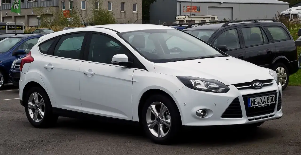 Biały Ford Focus MK3 1.6 benzyna zaparkowany obok innych samochodów.