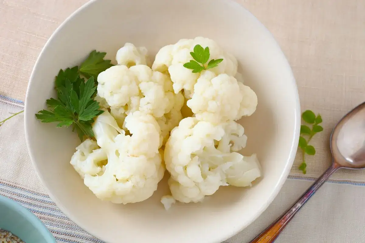 Cavolfiore bollito servito in una ciotola bianca, guarnito con prezzemolo fresco. Un cucchiaio d'argento è accanto.